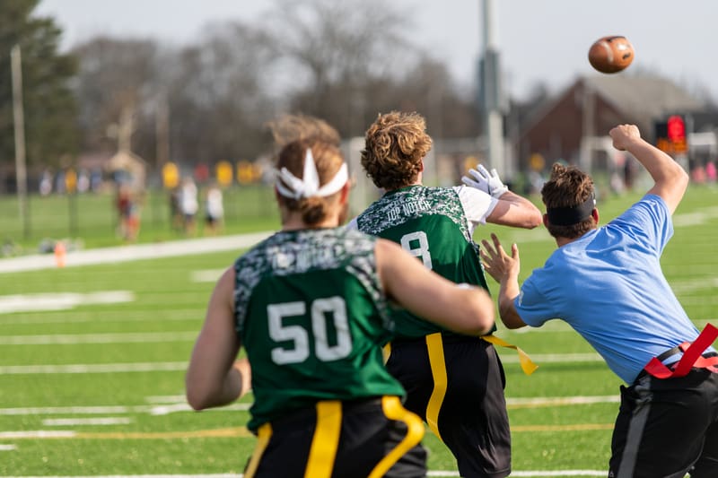 Three people play flag football on a grassy field. One in a blue shirt throws a football, while two in green jerseys run toward the end zone.